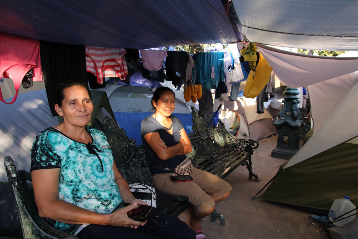 Two women sit in the shelter of tents, with clothing hanging on lines behind them.