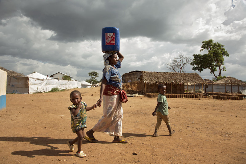 A woman walking with a jug of water on her head and two children next to her.
